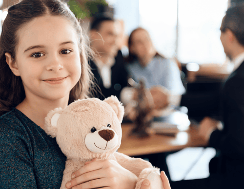 Young girl holding a teddy bear in one hand and smiling warmly.